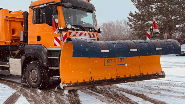 Ein orangefarbener Schneepflug steht auf einem mit Schnee bedeckten Weg. Der Lkw hat eine große, breite Schneeschaufel vorne und einen schwarzen Kasten auf der Rückseite. Im Hintergrund sind ein Gebäude und Bäume sichtbar.