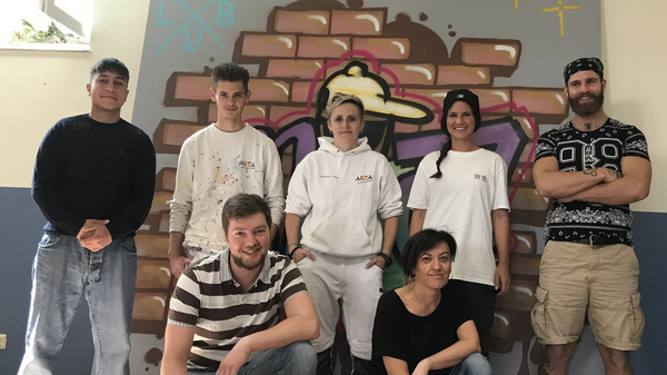 Eine Gruppe von sieben Personen steht vor einem Wandgemälde mit einer Mauer aus Ziegelsteinen und bunten grafischen Elementen. Die Teilnehmer sind unterschiedlich gekleidet und lächeln in die Kamera, während sie in zwei Reihen positioniert sind.
