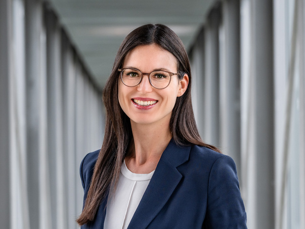 Eine lächelnde Frau mit langen, glatten Haaren trägt eine Brille und einen elegantenblauen Blazer. Sie steht in einem modernen Flur mit Glaswänden, der helle und gut beleuchtet ist.