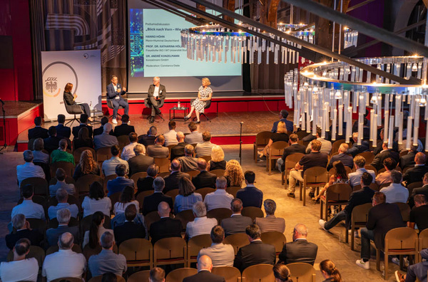 Eine Diskussion findet auf einer Bühne statt, unterstützt von einer Präsentation im Hintergrund. Vier Personen, zwei auf der Bühne und zwei im Publikum, sind in einem modernen Raum mit Stühlen versammelt. Das Publikum besteht aus vielen Zuhörern, die aufmerksam auf die Diskussion achten.