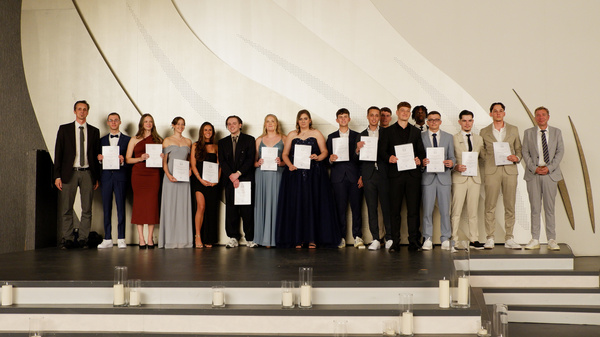 Eine Gruppe von 15 Personen steht auf einer Bühne, alle halten Urkunden in der Hand. Sie tragen festliche Kleidung und lächeln, während sie nebeneinander aufgereiht sind. Im Hintergrund ist eine dekorative Wand.