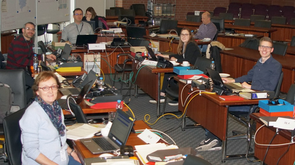 A group of diverse individuals is seated at desks in a large room, each using laptops and working on documents. There are multiple desks arranged with equipment and papers, suggesting a collaborative work environment. The setting appears professional and organized.