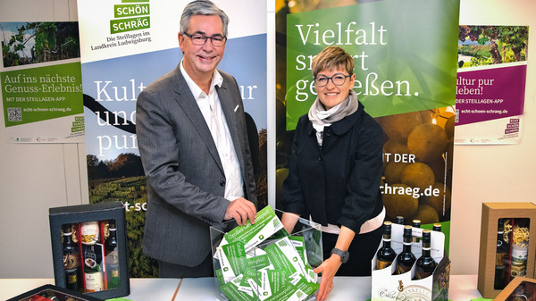 Zwei Personen stehen an einem Tisch, umgeben von Werbeflächen für regionale Produkte. Sie halten eine grüne Box mit dem Schriftzug „Vielfalt“. Auf dem Tisch liegen verschiedene Wein- und Likörflaschen sowie andere verpackte Produkte. Die Szene vermittelt ein Gefühl von lokaler Genussvielfalt.