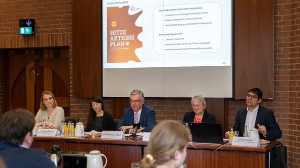 Eine Gruppe von fünf Personen sitzt an einem Tisch in einem Konferenzraum. Im Hintergrund ist eine Präsentation mit dem Titel "Hitzaktionsplan" zu sehen. Einige Getränke und Snacks stehen auf dem Tisch. Die Anwesenden diskutieren über wichtige Themen.