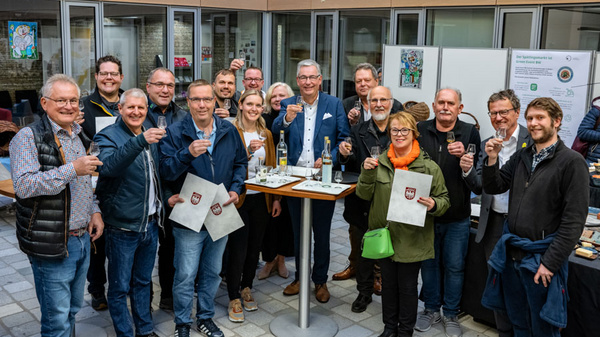 Eine Gruppe von etwa 20 Personen steht in einem Innenhof und hebt Gläser zum Anstoßen. Sie lächeln und tragen unterschiedliche Kleidung. Im Hintergrund sind Informationsstände und farbige Kunstwerke zu sehen. Die Szene vermittelt festliche Stimmung und Gemeinschaftlichkeit.