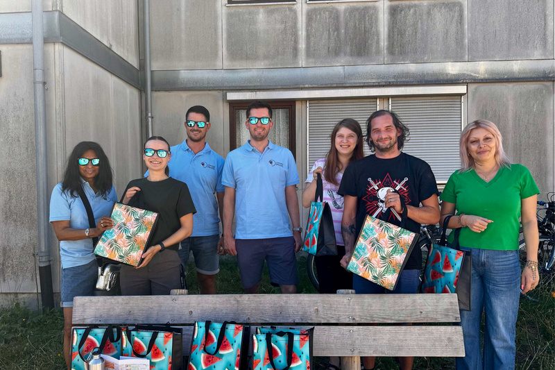 Eine Gruppe von acht Personen steht vor einem Gebäude und hält farbenfrohe Taschen mit Wassermelonen-Motiv. Alle tragen Sonnenbrillen und lächeln. Im Vordergrund sind einige gesunde Snacks auf einer Bank sichtbar.