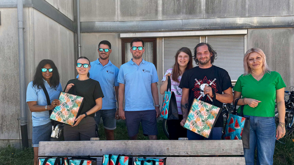 Eine Gruppe von acht Personen steht vor einem Gebäude und hält farbenfrohe Taschen mit Wassermelonen-Motiv. Alle tragen Sonnenbrillen und lächeln. Im Vordergrund sind einige gesunde Snacks auf einer Bank sichtbar. (Text KI generiert)
