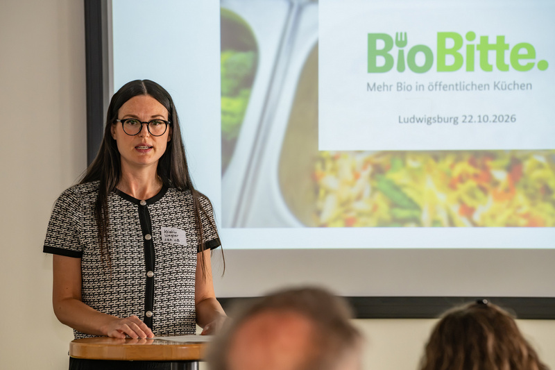 Eine Frau steht an einem Podium und spricht vor einem Bildschirm, auf dem das Logo von "BioBitte" und Informationen über eine Veranstaltung in Ludwigsburg angezeigt werden. Sie trägt eine Schwarz-Weiß-Oberbekleidung und hat lange, dunkle Haare. (Text KI generiert)