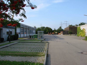 Eine breite, gepflasterte Straße führt durch ein industrielles Gebiet mit mehreren Gebäuden auf beiden Seiten. Im Vordergrund steht eine Bushaltestelle. Bäume und Sträucher sind in der Umgebung sichtbar. Der Himmel ist klar und blau, was eine ruhige Atmosphäre vermittelt.