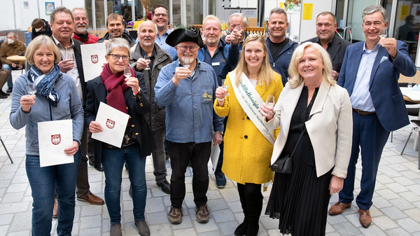 Groupe de personnes se réunissant pour célébrer un événement. Plusieurs participants tiennent des verres en l'air pour porter un toast, tandis que d'autres montrent des documents. Une femme portant une couronne et une écharpe se distingue au centre. L'atmosphère est festive et conviviale.