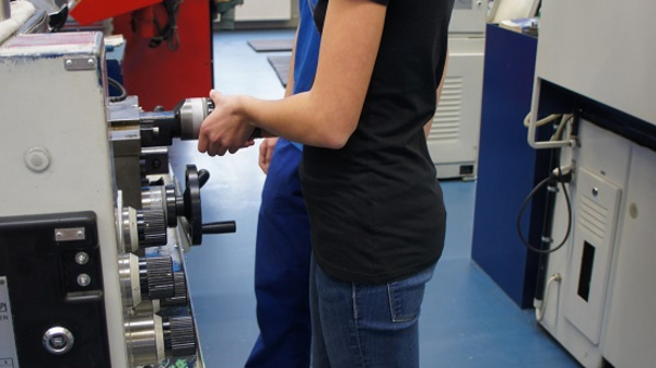 Eine junge Frau steht an einer Maschine in einer Werkstatt und bedient diese. Sie trägt ein schwarzes T-Shirt und eine Schutzbrille. Im Hintergrund ist eine andere Person zu sehen, die ebenfalls an der Maschine arbeitet.
