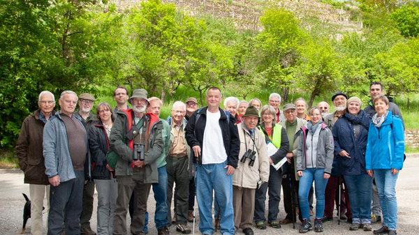 A large group of diverse adults is standing together outdoors in a green, forested area. They appear to be participants in a nature or hiking event, dressed in casual attire, with some holding binoculars and wearing hats. The atmosphere is cheerful and engaged.