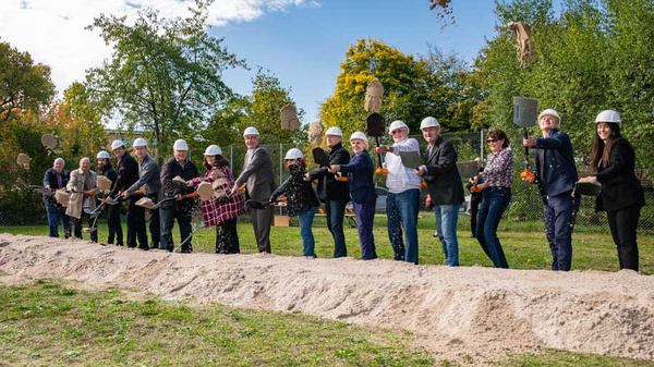 Eine Gruppe von Menschen in Bauhelmen steht an einer Erdgrube und hält Schaufeln. Sie posieren gemeinsam für ein Foto, während einige von ihnen Erde in die Luft werfen. Die Szene deutet auf einen Spatenstich oder Baubeginn hin. Es ist ein sonniger Tag mit grüner Landschaft im Hintergrund.