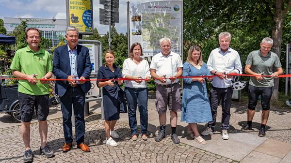 Otto persone, tra cui uomini e donne, stanno partecipando a un'inaugurazione, tenendo in mano un nastro rosso da tagliare. Si trovano all'esterno, in un'area ciottolata, con un cartello informativo sullo sfondo. L'atmosfera è festosa e celebrativa.