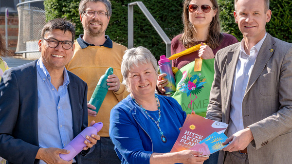 Sechs Personen stehen auf einer Treppe und halten verschiedene bunte Gegenstände, darunter Trinkflaschen und Plakate. Sie lächeln und scheinen eine positive Botschaft zu präsentieren. Im Hintergrund sind grüne Sträucher und ein modernes Gebäude zu sehen.