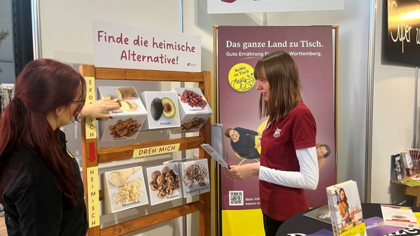 Zwei Frauen stehen am Informationsstand des Ernährungszentrums Mittlerer Neckar. Eine Frau zeigt auf eine Tafel mit Bildern von Lebensmitteln und dem Text „Finde die heimische Alternative!“ Im Hintergrund ein Info-Plakat über regionale Ernährung.