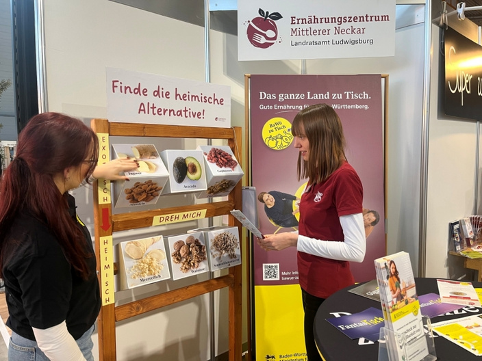 Zwei Frauen stehen am Informationsstand des Ernährungszentrums Mittlerer Neckar. Eine Frau zeigt auf eine Tafel mit Bildern von Lebensmitteln und dem Text „Finde die heimische Alternative!“ Im Hintergrund ein Info-Plakat über regionale Ernährung.