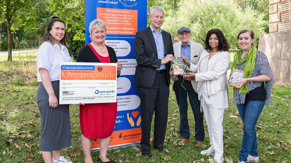 Eine Gruppe von sechs Personen steht im Freien und hält einen großen Scheck zur Organspende. Im Hintergrund ist ein Banner mit Informationen zur Organspende sichtbar. Alle Personen wirken freundlich und engagiert in ihrer Unterstützung für die Organspendebewegung.
