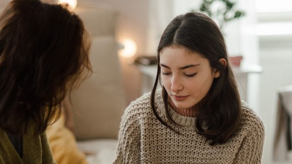 Zwei Frauen sitzen zusammen auf einem Bett. Eine Frau hält die Hände der anderen und sieht sie aufmerksam an. Die Atmosphäre ist unterstützend und einfühlsam, was auf eine vertrauliche Unterhaltung hindeutet.