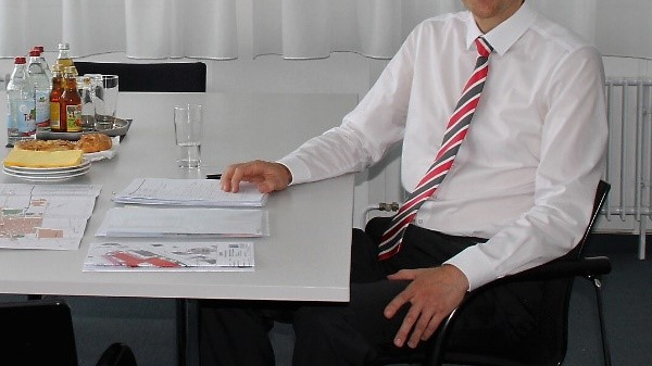 Two men are seated at a table in a bright office. They are smiling and engaged in conversation, with documents and maps spread on the table. A few bottles and cups are also present, suggesting a meeting or discussion taking place.