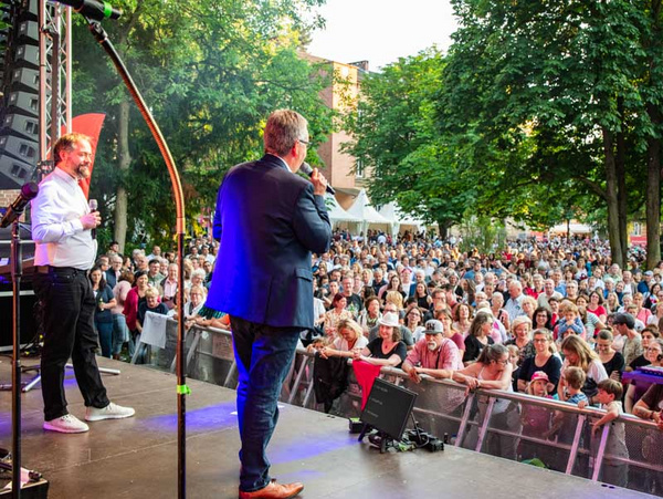 23.06.2023 - Begrüßung der Gäste des Landkreisfeschds Zwei Männer stehen auf einer Bühne und sprechen zu einem großen, begeisterten Publikum. Die Zuschauer stehen in Gruppen und scheinen der Darbietung aufmerksam zuzuhören. Bäume und Stände im Hintergrund schaffen eine festliche Atmosphäre.
