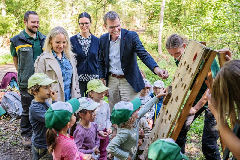 Eine Gruppe von Erwachsenen und Kindern steht im Wald um ein interaktives Lernspiel. Die Kinder, die grüne Caps tragen, sind begeistert und schauen auf das Spielbrett, während die Erwachsenen lächeln und mitdenkende Gesten machen. Der Hintergrund zeigt grüne Bäume. (Text KI generiert)