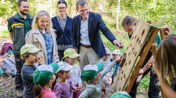 Eine Gruppe von Erwachsenen und Kindern steht im Wald um ein interaktives Lernspiel. Die Kinder, die grüne Caps tragen, sind begeistert und schauen auf das Spielbrett, während die Erwachsenen lächeln und mitdenkende Gesten machen. Der Hintergrund zeigt grüne Bäume. (Text KI generiert)