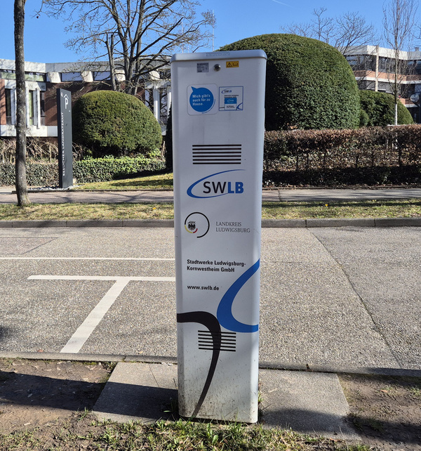 Ein weißer Informationsstand des SWLB steht an einer Straße, umgeben von Bäumen und Hecken. Der Stand zeigt das Logo der SWLB und enthält Informationen zu öffentlichen Dienstleistungen. Im Hintergrund sind parkende Autos und Gebäude sichtbar.