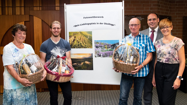A group of four people stands in front of a poster for a photography competition labeled “My Favorite Place in the Steillages.” Two participants hold decorated baskets filled with goods, while the others, dressed in professional attire, smile and pose for the photo.