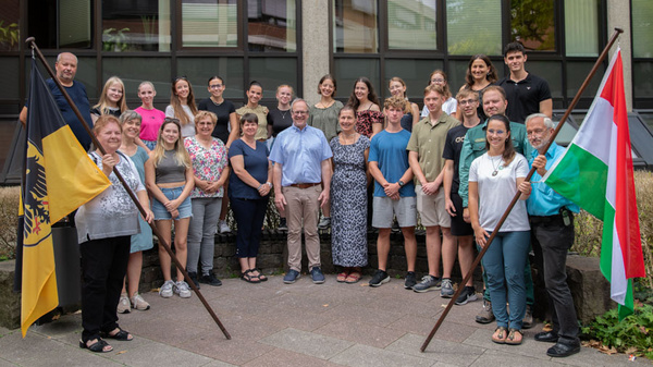 Eine Gruppe von etwa 30 Personen steht vor einem Gebäude. Einige halten Fahnen in den Händen. Die Gruppe besteht aus Menschen unterschiedlichen Alters, darunter Jugendliche und Erwachsene, die in einer freundlichen Pose zusammenstehen.