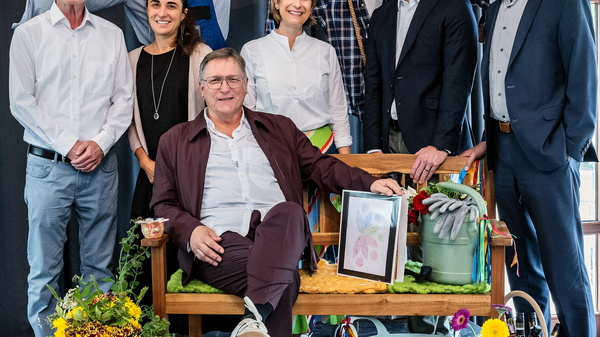 Eine Gruppe von sechs Personen steht und sitzt auf einer Bank vor einem schwarzen Vorhang. Die Männer und Frauen lächeln freundlich in die Kamera. Im Hintergrund sind zwei Figuren in Kostümen zu sehen. Auf der Bank sind Blumen und Gartenutensilien dekoriert.