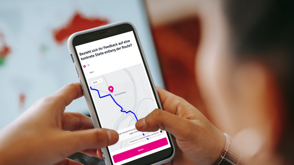 Due mani tengono uno smartphone che mostra una mappa con un percorso evidenziato. Il dispositivo è posizionato vicino a un computer, mentre sullo schermo del computer sono visibili grafici e mappe.