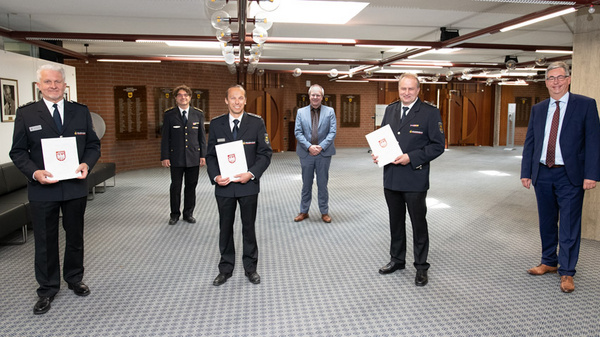 A group of five men stands together indoors, with four in police uniforms holding certificates and one in a suit. They appear to be celebrating an achievement or recognition, with a few smiling and others standing proudly in a spacious, well-lit area.