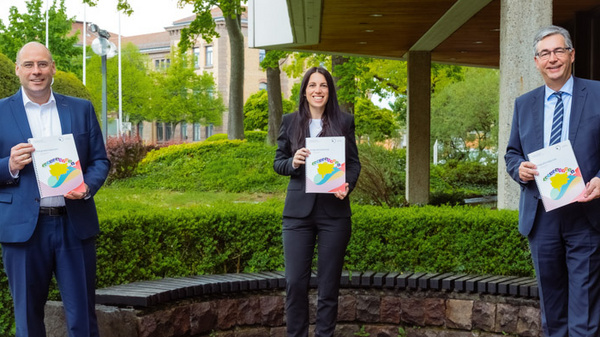 Three professionals stand outside a building, holding documents featuring a colorful map design. The scene is surrounded by greenery, indicating a pleasant environment. They appear to be engaged in a positive discussion or event, highlighting the importance of the document they are presenting.