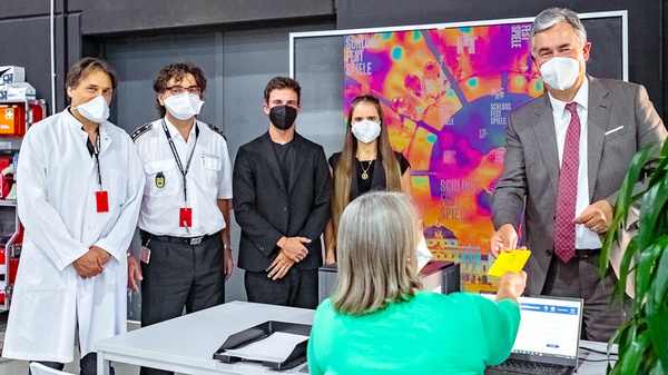 A group of five people stands in a modern indoor setting. They are wearing masks due to health guidelines. One person in the foreground, wearing a green shirt, is handing a yellow piece of paper to a man in a suit. The background features colorful digital artwork.