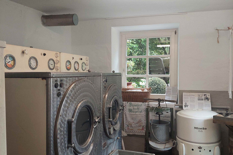 Das Bild zeigt einen Raum mit zwei großen Waschmaschinen an der Wand, die mit Druckmessern ausgestattet sind. Neben den Waschmaschinen steht ein Wäschewagen. Durch ein Fenster ist Außenlicht sichtbar, und an der Wand hängen einige Dokumente.