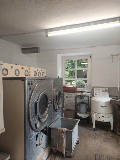 Das Bild zeigt einen Raum mit zwei großen Waschmaschinen an der Wand, die mit Druckmessern ausgestattet sind. Neben den Waschmaschinen steht ein Wäschewagen. Durch ein Fenster ist Außenlicht sichtbar, und an der Wand hängen einige Dokumente.