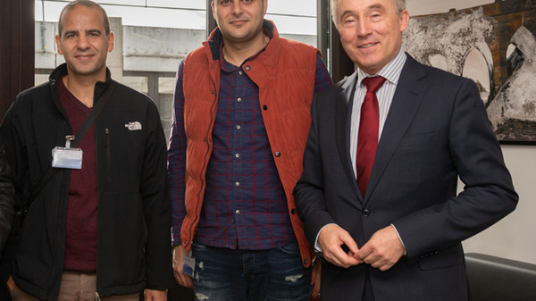 Four men are standing together in an indoor setting. They are smiling and dressed in casual and formal attire. Large windows in the background suggest a bright, spacious environment. The overall mood is friendly and professional, indicating a meeting or collaborative event.