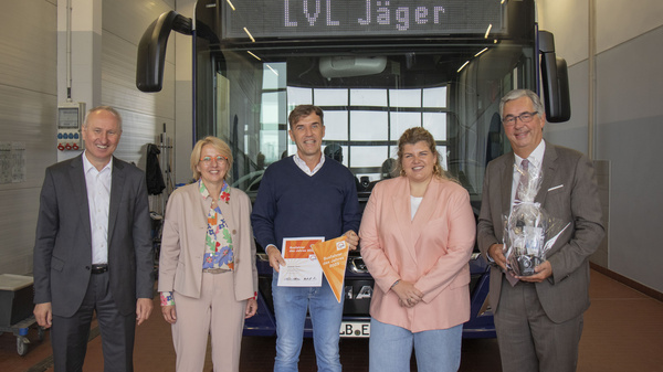 Fünf Personen stehen vor einem Bus mit der Aufschrift „LVL Jäger“. Sie lächeln und halten Urkunden oder Preise in den Händen. Die Szene zeigt eine feierliche Übergabe oder Auszeichnung im Transportbereich.