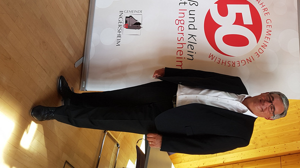 Zwei Personen stehen vor einem Banner zum 50-jährigen Jubiläum einer Gemeinde. Das Banner zeigt die Zahl „50“ und den Text: „Groß und klein ist ein bisschen Community“. Der Hintergrund ist aus hellem Holz, und die Umgebung wirkt freundlich und einladend.