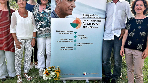 A group of seven people stands together outdoors near a banner. The banner features an image of an older man and text in German promoting support for individuals diagnosed with dementia, emphasizing the importance of staying involved and connected after diagnosis.