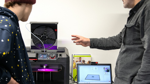 Ein älterer Mann und ein Jugendlicher stehen neben einem 3D-Drucker. Der Mann erklärt etwas, während der Jugendliche aufmerksam zuhört. Auf einem Laptop neben dem Drucker sind Grafiken und Informationen sichtbar. Die Szene vermittelt eine Lehrsituation über die Verwendung von 3D-Drucktechnologie.
