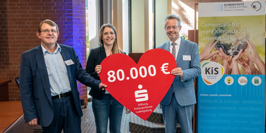 Drei Personen stehen in einem Raum und halten ein großes rotes Herz mit der Aufschrift "80.000 €". Im Hintergrund ist ein Banner zu sehen, das über Klimaschutz in Schulen informiert. Die Personen wirken zufrieden und engagiert für das Thema.
