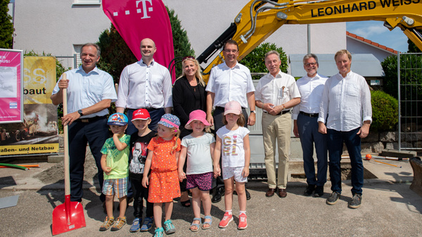 A group of officials and children gather at a construction site. Several adults, including some with shovels, stand behind young children dressed in summer clothing. A construction vehicle is partially visible in the background, indicating an event related to building or infrastructure development.