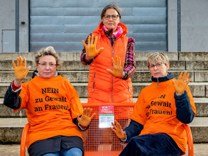 Tre donne in t-shirt arancioni con la scritta "NEIN zu Gewalt an Frauen!" siedono su una panchina. Hanno le mani dipinte di arancione e sollevano le mani per evidenziare il loro messaggio di protesta contro la violenza sulle donne.