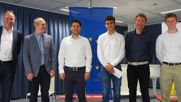 Sechs Männer stehen in einer Reihe in einem Konferenzraum. Sie tragen verschiedene Formate eleganter Kleidung und lächeln. Im Hintergrund ist eine EU-Flagge zu sehen. Der Raum wirkt professionell und festlich, passend zu einer besonderen Veranstaltung.
