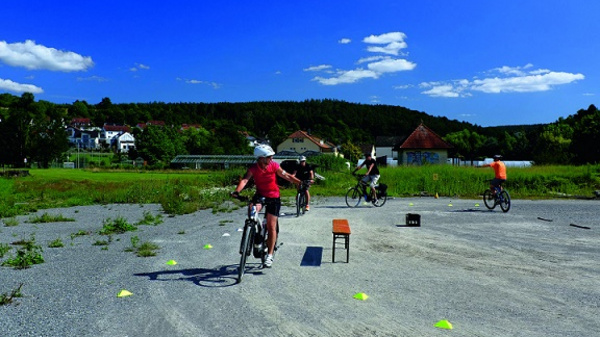 Des enfants pratiquent le cyclisme sur un terrain dégagé. Ils montent des vélos, passant autour de cônes posés sur le sol. Le ciel est bleu, avec quelques nuages, et la campagne est visible en arrière-plan.