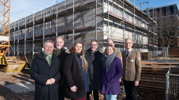 A group of seven people poses in front of a partially constructed building surrounded by scaffolding. They are standing together, smiling, on a clear day. The scene includes a yellow construction crane on the left.
