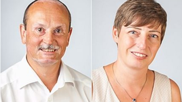 Das Bild zeigt zwei Personen: einen Mann mit Schnurrbart und einem weißen Hemd links, und eine Frau mit kurzem, braunem Haar und einem hellen Oberteil rechts. Beide lächeln freundlich in die Kamera vor einem neutralen Hintergrund.