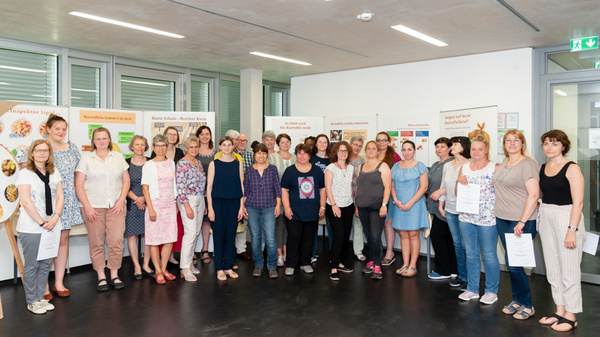 A diverse group of around 30 people stands together in a well-lit room, posing for a photo. They appear engaged and are holding documents. Behind them are presentation boards displaying various topics, indicating a gathering centered around education or community involvement.